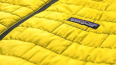 노란색 퀼팅 처리된 파타고니아(Patagonia) 재킷의 근접 사진. 검은색 지퍼와 가슴 부분의 파타고니아 로고 태그가 선명하게 보인다. B-Corp 인증 기업의 친환경 제품 철학을 보여주는 이미지.