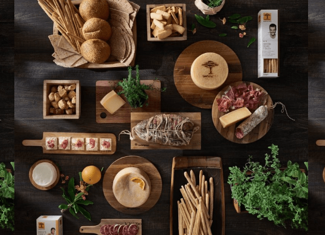 어두운 나무 질감의 배경 위에 산 파트리냐노(San Patrignano) 공동체의 로고가 선명하게 새겨진 치즈, 가공육(살라미/햄), 빵, 신선한 허브 등 고품질 식품들이 목재 트레이에 감각적으로 배치된 모습. 사회적 협동조합의 전문적인 직업 훈련과 경제적 자립을 상징하는 이미지.