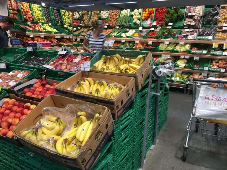 푸른 하늘과 눈 덮인 산이 보이는 스위스 체르마트(Zermatt)의 전통적인 목조 건축물들 사이에 위치한 스위스 쿱(Coop) 매장 전경. 현대적인 로고와 지역 전통 양식이 조화를 이룬 유통 매장의 모습.