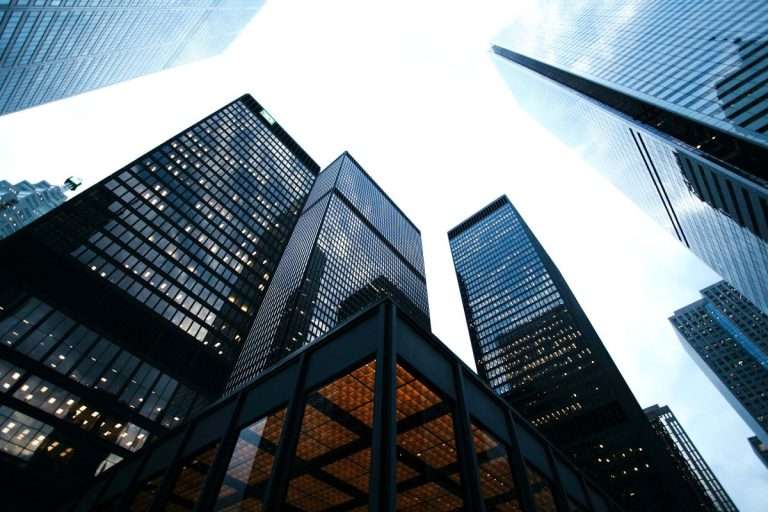 수직으로 높게 솟은 현대적인 유리 외벽 빌딩들을 아래에서 위로 올려다본 모습. 도심 마천루의 기하학적인 구조와 투명한 창들이 어우러진 전문적이고 견고한 기업 환경 이미지.