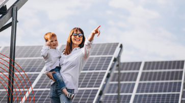 푸른 하늘 아래 설치된 대규모 태양광 패널 단지 앞에서 선글라스를 쓴 여성이 어린아이를 안고 한 손으로 먼 곳을 가리키며 밝게 웃고 있는 모습. 재생에너지로의 전환과 지속가능한 미래를 상징하는 사진.