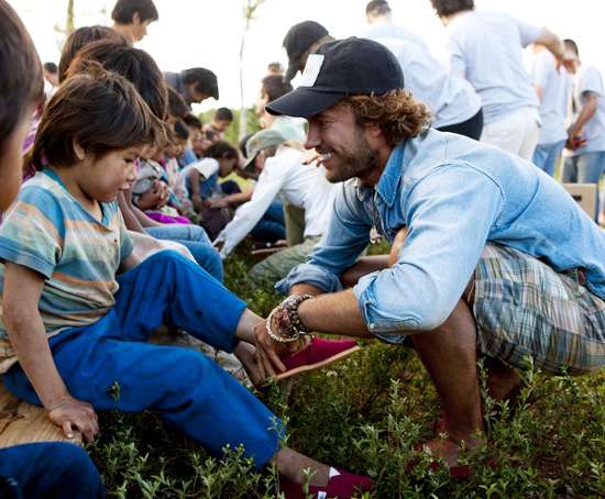 탐스 슈즈의 설립자 블레이크 마이코스키가 먼지가 날리는 야외에서 어린아이의 발에 직접 신발을 신겨주며 소통하는 모습. 원포원(One for One) 기부 모델의 실천 현장을 상징하는 사진.