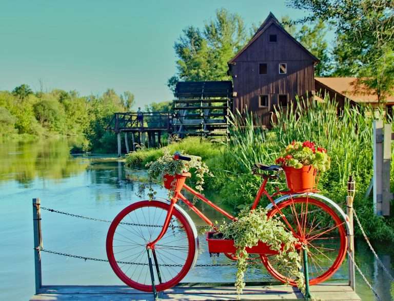 강변 데크 위에 놓인 꽃바구니가 달린 빨간 자전거와 배경으로 보이는 강물, 울창한 나무, 그리고 전통적인 목조 물레방아 건물이 어우러진 평화로운 자연 풍경