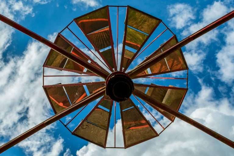 푸른 하늘과 흰 구름을 배경으로 아래에서 위로 올려다본 원형의 금속 구조물. 자전거 바퀴나 풍차를 연상시키는 기하학적인 패턴이 자원의 끊임없는 회전과 선순환을 상징하는 이미지.