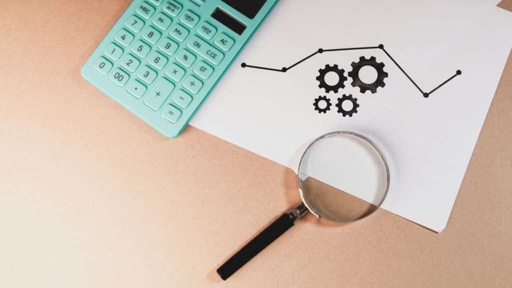 계산기, 돋보기, 그리고 톱니바퀴와 그래프가 그려진 문서가 책상 위에 놓여 있는 이미지. 데이터 기반의 정밀한 비즈니스 분석과 프로세스 진단 과정을 상징합니다.